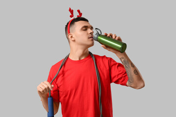 Sticker - Young man in reindeer horns with skipping rope drinking water on grey background