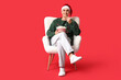© Pixel-Shot - Young man with bucket of popcorn watching movie and sitting in armchair on red background. Christmas celebration