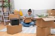 © olga_demina - Embracing Change: A young woman in her new home, surrounded by carton boxes, unfolds her belongings, a symbol of relocation, rentals, or home buying