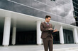 © dikushin - Handsome businessman in suit professional working on laptop standing outside modern office building, exuding confidence and determination in urban setting. Concept of business communication.