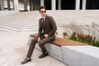 © dikushin - Full length portrait of of stylish photogenic young businessman in brown suit and sunglasses sitting confidently on city bench, exuding professionalism and success in modern urban setting.