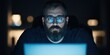 © Kamonwan - Focused man working late at night with laptop, illuminated by screen light, serious expression, dark setting.