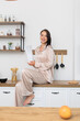 © Євген Хоптинський - Smiling woman in cozy loungewear sitting on a kitchen counter, holding a container. Modern, bright kitchen setting with organized shelves and fresh ingredients, embodying a relaxed lifestyle.