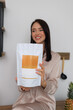© Євген Хоптинський - A smiling woman in soft loungewear holds a white packaging pouch with a yellow label in a cozy kitchen setting. Perfect for promoting healthy products, supplements, or food packaging concepts.