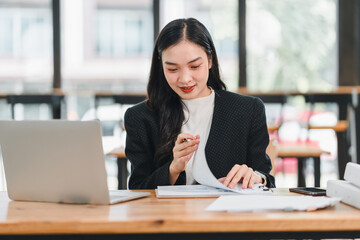 Wall Mural - young woman in black blazer is working at desk with laptop, reviewing documents and taking notes. She appears focused and engaged in her task