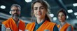 © Riko - Portrait of confident female engineer in orange safety vest with colleagues in a factory background