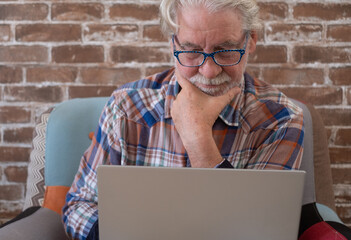 Wall Mural - Smiling attractive senior man sitting at home on armchair using laptop computer. Brick wall in background