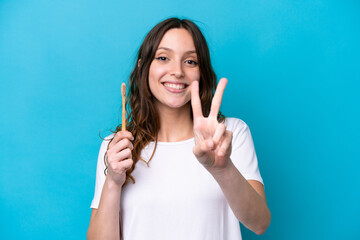 Wall Mural - Young caucasian woman brushing teeth isolated on blue background smiling and showing victory sign