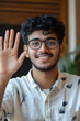 © Prasanth - Cheerful positive young Indian student guy in glasses waving hand hello at camera, making greeting gesture on video call, speaking on conference chat, smiling. Vertical head shot portrait