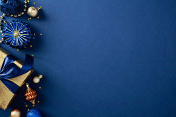 Wall Mural - Stylish Christmas ornaments in gold and blue on a dark blue background. Includes glittery baubles, gold gift box with blue ribbon bow, confetti. Flat lay, top view, copy space