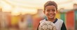 © Naphol - A cheerful young boy smiles while holding a soccer ball outdoors, showcasing joy and enthusiasm for the game during sunset.