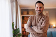 © Moon Safari - Portrait of positive handsome man with arms crossed smiling at camera and standing by window at home