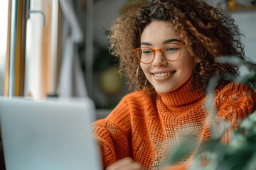happy woman wearing glasses and orange sweater using laptop sitting at desk taking notes watching on