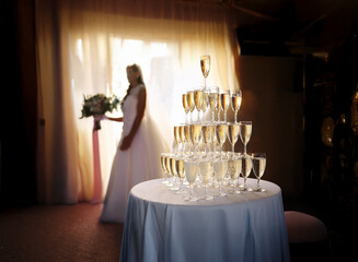 A bride in a white wedding dress walks towards a champagne pyramid on a white table.