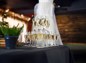 A pyramid of champagne glasses sits on a black tablecloth with a plant in a gray pot and a white dress in the background.