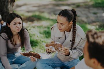 Poster - A group of young students sitting together on grass, enjoying sandwiches under the shade of a tree in a park. They appear relaxed and happy in the warm outdoor setting.