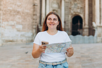 Wall Mural - Attractive female tourist is exploring new city. Redhead woman holding a paper map near the Virgin Saint Mary, Valencia Cathedral. Traveling Europe