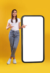 Wall Mural - Mobile App Advertisement. Full Body Length Of Happy Woman Showing Pointing At Big Huge White Empty Smartphone Screen Standing Over Orange Studio Background. Check This Out, Cellphone Display Mock Up