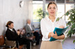 © JackF - Smiling young female business assistant standing in office with clipboard in hands, noting tasks