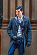 © Alexander Image - A well-dressed man stands confidently in front of a stone column, wearing a dark overcoat and gloves with a crossbody bag. His poised stance and stylish attire suggest a sense of urban sophistication.