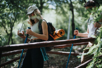 Wall Mural - Couple enjoying a hike in the forest with camping gear. Woman drinking iced coffee while holding a walking stick.