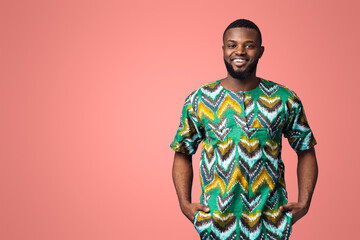 Wall Mural - Handsome black man in traditional african clothes smiling over blue studio background, putting hands in pockets, copy space. Cheerful african american guy posing in ethnic colorful clothes