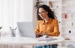 © Prostock-studio - Middle Eastern Lady Using Laptop Working Online Wearing Eyewear Sitting At Workplace In Modern Office. Remote Job, Technology And Career Profession Concept. Side View