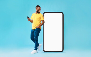 Wall Mural - cheery millennial black guy with mobile device looking at giant smartphone on blue background, mocku