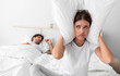 © Prostock-studio - Despaired millennial european wife sits on bed covers ears with pillow and suffers from snoring of sleeping husband in bedroom interior. Intolerable snoring and noise at night, insomnia and apnea