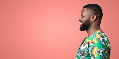 Wall Mural - Side view of black smiling guy in african shirt looking at free space on blue, studio shot, panorama. Excited african american young man staring at great deal or offer over colorful background