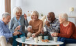 © Prostock-studio - Multiethnic group of happy senior friends men and women in casual sitting on couch, having conversation and playing cards while drinking tea with cookies, chilling together at home