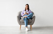 © Prostock-studio - Beautiful young woman sitting in armchair, looking at mobile phone screen, using new cool application against white studio wall, full length. Joyful lady using smartphone for work or entertainment