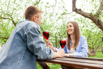 Wall Mural - young couple in love in denim clothes sitting in the garden at a wooden table and drinking red wine with grapes on a date in the summer, guy and girl trying alcoholic cocktails and flirting in the