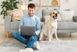 © Prostock-studio - Working Home Concept. Portrait of smiling man sitting on carpet in the living room with his labrador, using laptop computer. Happy guy doing freelance job in internet online, teleworking on the floor