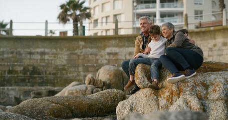 Wall Mural - Grandparents, child and happy on rocks at beach with hug, memory and outdoor on holiday with bonding. Senior man, woman and boy with embrace, care and love with family on vacation with view in Italy