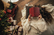 © polinaloves - Young woman reading book, lying near Christmas tree in a cozy decorated room.