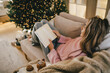 © polinaloves - Young woman relaxing, reading book and drinking cocoa in a cozy living room near Christmas tree.