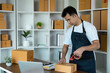 © itchaznong - Young Man in Apron Packing Boxes for Shipping in Home Office with Laptop and Supplies on Desk