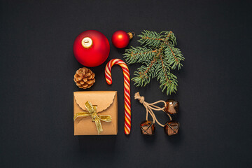 Festive flat lay features red and craft ornaments, candy cane, pine branch and gift box, all arranged on black fabric background, evoking spirit of holiday season.