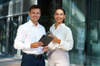 © muse studio - Two Professionals Standing Outside a Modern Office Building, Engaging With Digital Devices on a Sunny Day