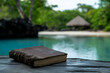 © Rohit k  - A weathered book rests on a wooden table near tranquil waters surrounded by lush greenery and a thatched-roof hut in the background, inviting contemplation in a serene setting