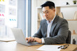 © sofiko14 - Asian businessman working on laptop in modern office setting. Young professional wearing suit focused on computer tasks. Office environment with bookshelf and documents on desk.
