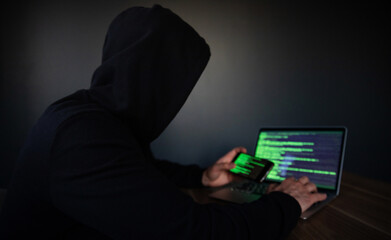 A hooded person operates a laptop and smartphone displaying green code, symbolizing hacking, cybercrime, or cybersecurity. The dark background enhances the secretive, tech-focused atmosphere.