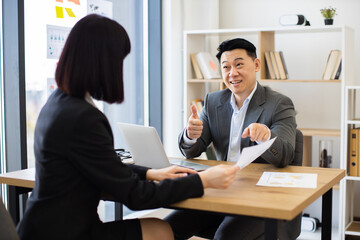 Wall Mural - Asian businessman discusses report with Caucasian businesswoman in modern office. Professional setting includes laptops, charts, and documents.