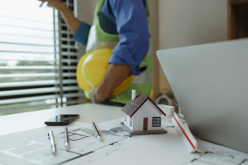 Poster - there are scientific calculator, laptop, house model, blueprint on the desk in the office with two architects, engineer is meeting with contractor about instruction project