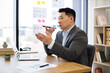 © sofiko14 - Asian male businessman in modern office sending audio messages via smartphone. He appears focused, demonstrating an engaging work environment. Office features laptop, documents, organized setting.