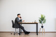 © sofiko14 - Asian man in formal suit working on laptop while making business call at desk. Modern workspace features light decor with white wall and plant. Represents concepts of communication, work, focus.