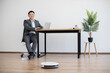 © sofiko14 - Asian businessman in suit works on laptop at modern office desk. Robot vacuum cleaner operates on wooden floor providing efficient cleaning. Bright workspace includes potted plant minimalist furniture