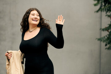 Brightly smiling, a young woman in a black dress waves warmly while enjoying the autumn air outdoors.