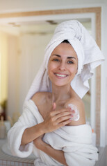 Wall Mural - A smiling woman in a white bathrobe and towel applies lotion to her shoulder in a bright bathroom, emphasizing skincare, self-care, and relaxation. Perfect for promoting beauty or wellness products.